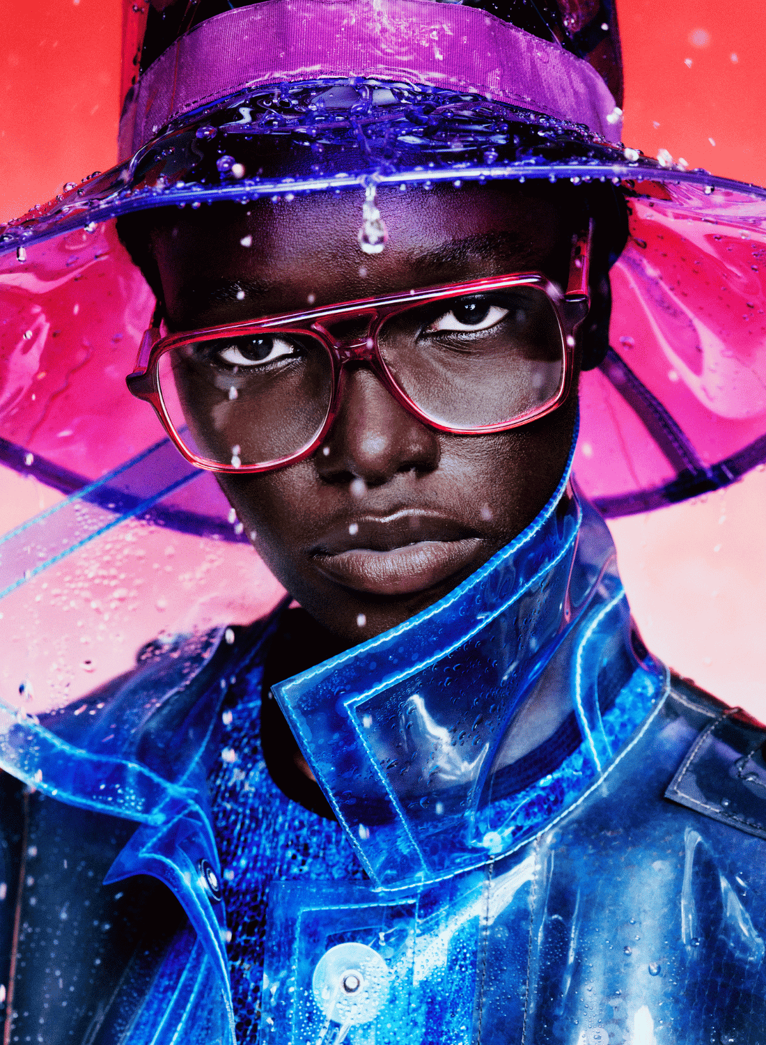 Person in a glossy blue coat and pink hat, wearing red glasses against a vibrant pink background