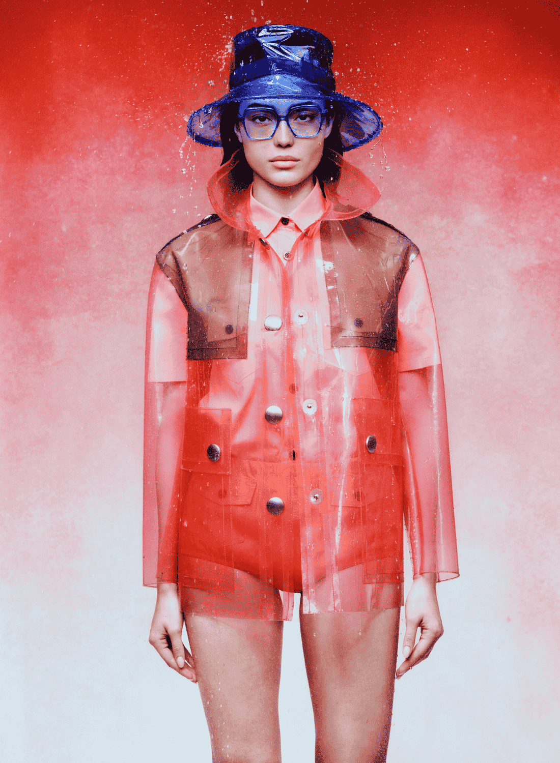 Model in a translucent red raincoat and blue hat against a red background.