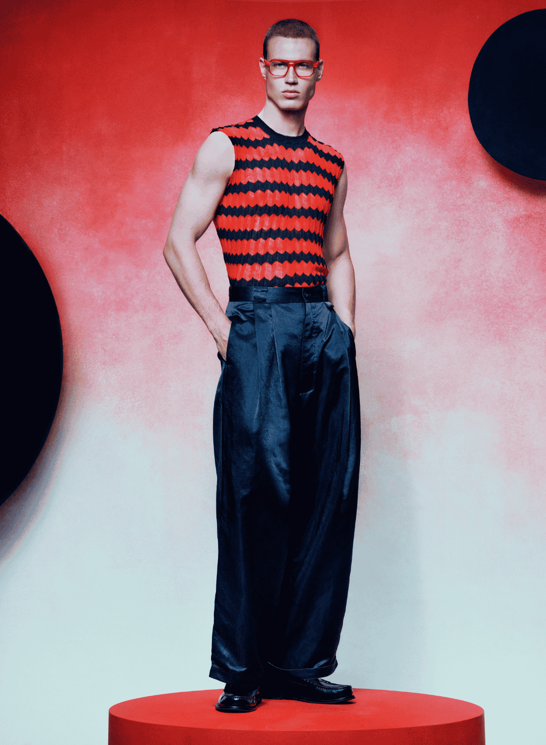 Model in red-and-black striped top and dark pants standing on a red pedestal against a red backdrop.