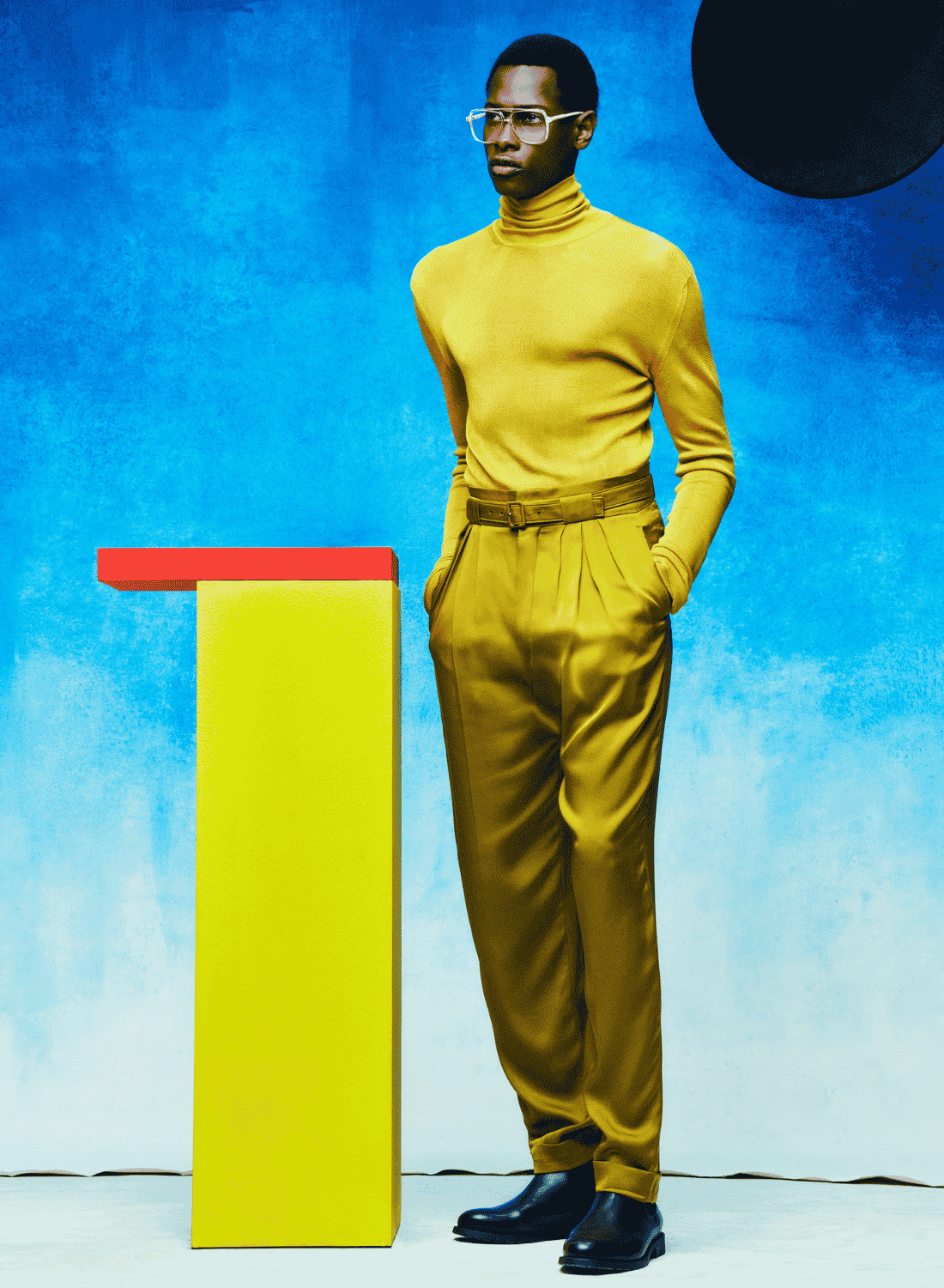 Man in mustard turtleneck and pants stands beside a yellow pedestal with a red top on a blue backdrop.