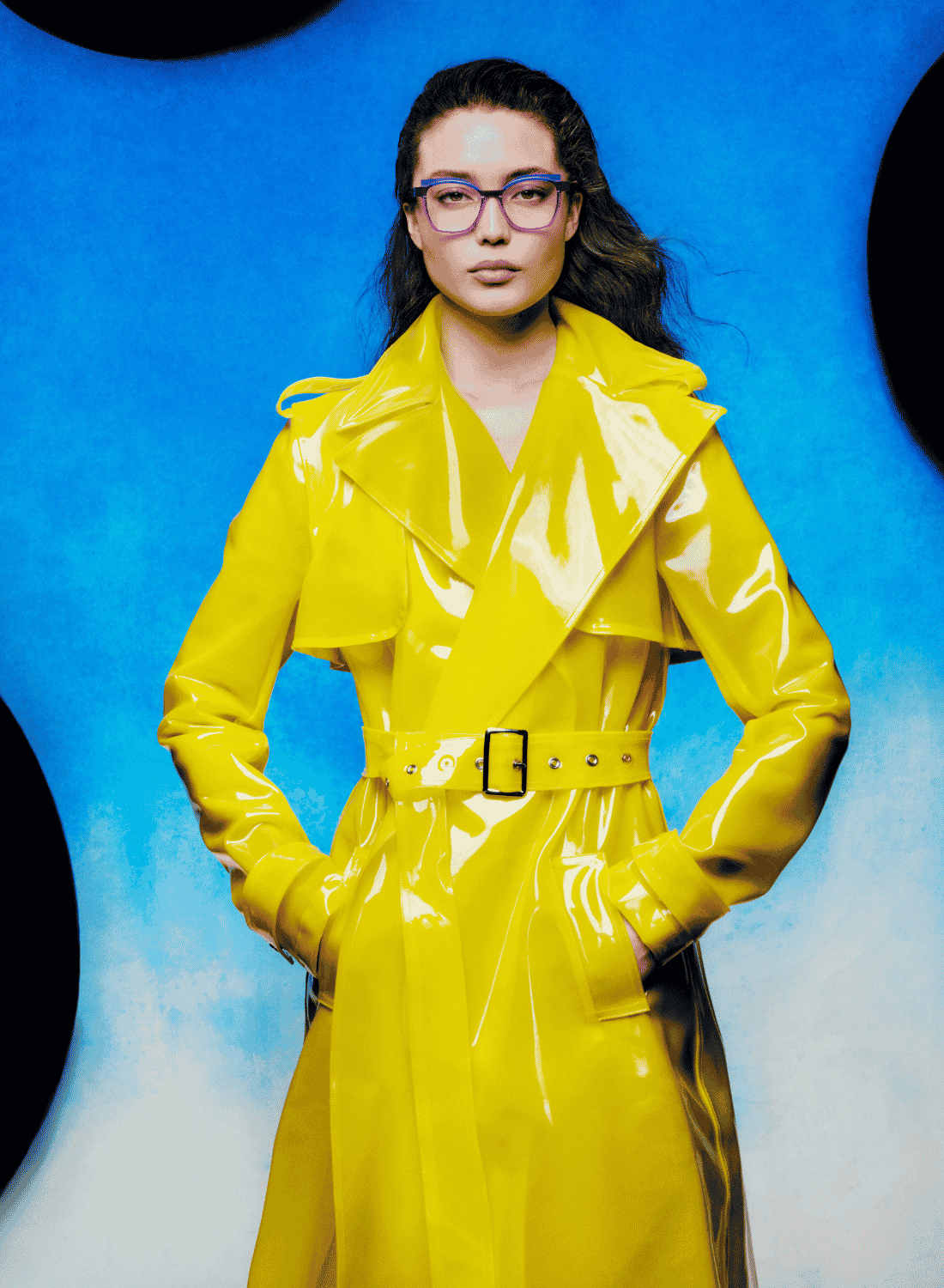 Person in a bright yellow trench coat and glasses stands against a blue background.