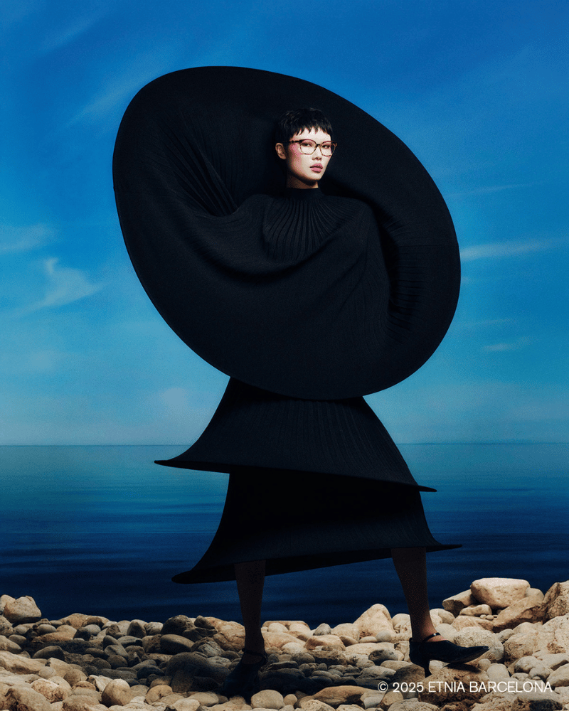 A woman wearing glasses in a black dress is standing on rocks near the ocean