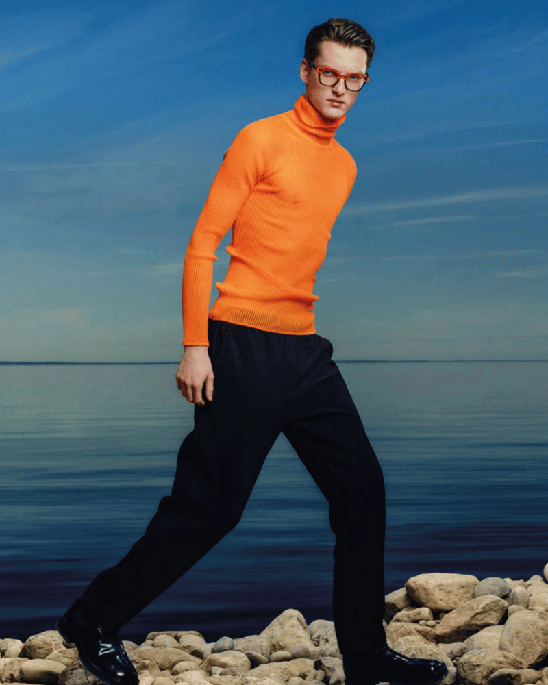 A man wearing glasses, an orange turtleneck sweater and black pants is walking on a rocky beach.