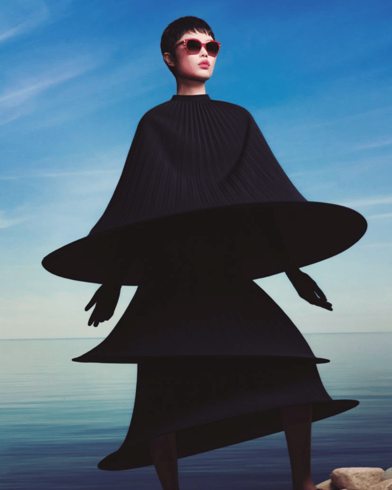 A woman wearing sunglasses and a black dress is standing in front of the ocean