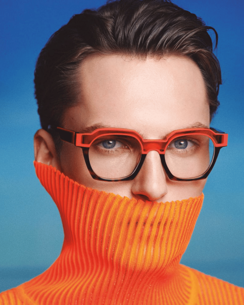 A man wearing glasses and an orange turtleneck covering his face