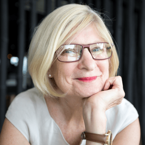 Woman with blonde hair and glasses, resting chin on hand, smiling.
