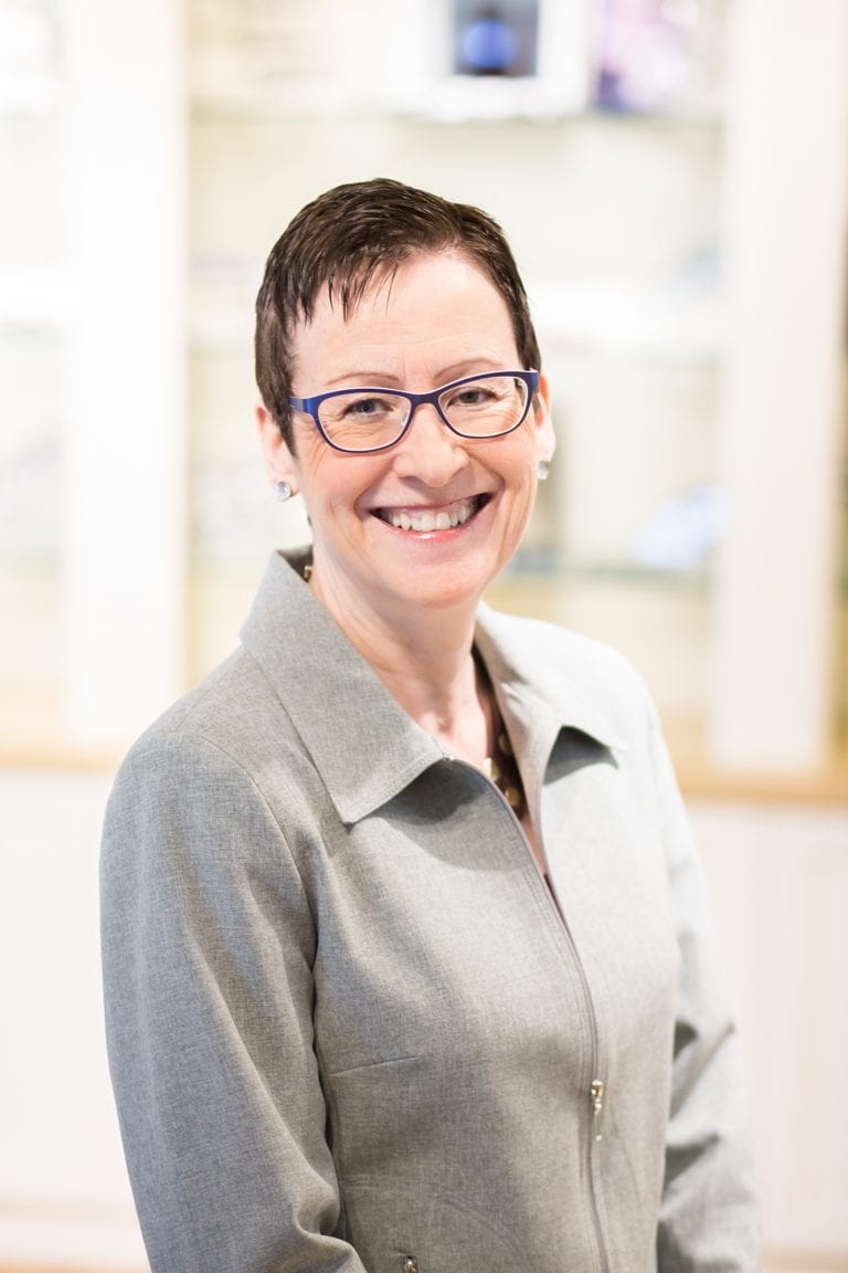 A woman wearing glasses and a grey jacket is smiling for the camera.