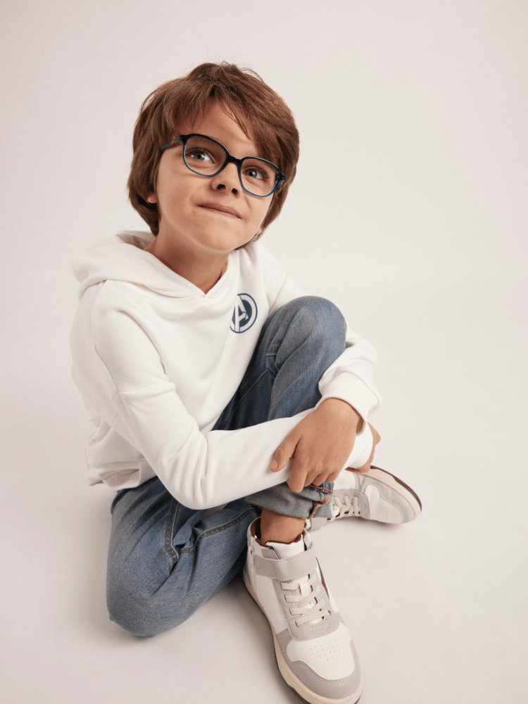 A young boy wearing glasses and a white hoodie is sitting on the floor.