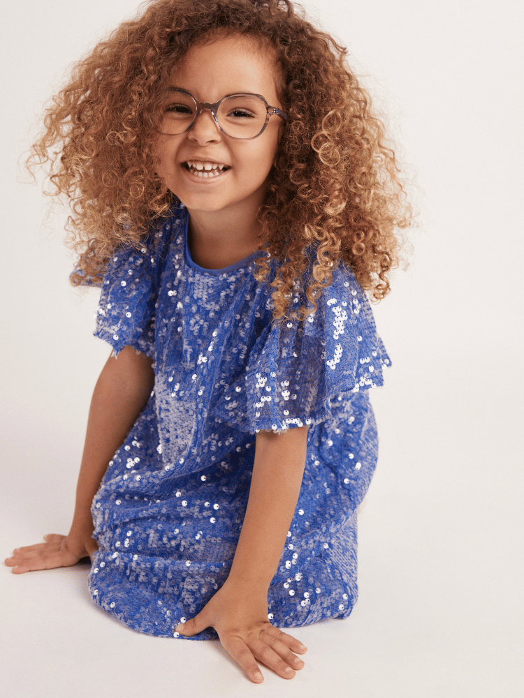 A little girl wearing glasses and a blue sequined dress is sitting on the floor.