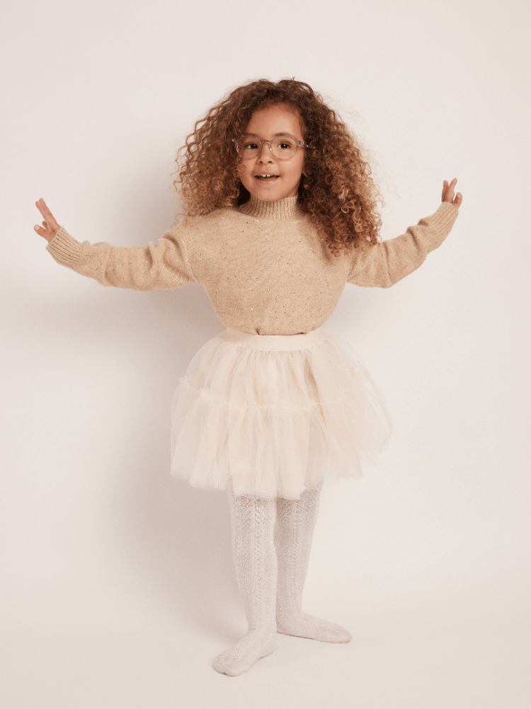 A little girl wearing glasses, a sweater and a white tulle skirt is standing with her arms outstretched.