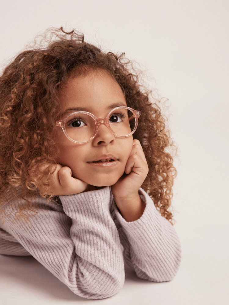 A little girl wearing glasses and a sweater is laying on the floor.