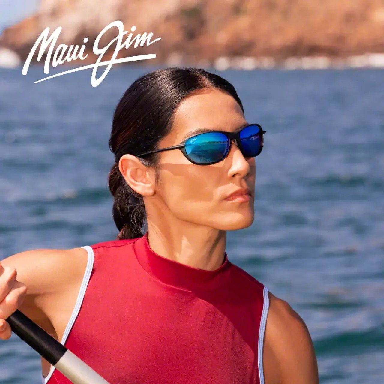 Woman in red top and blue sunglasses paddling on calm water with Maui Jim logo in the corner