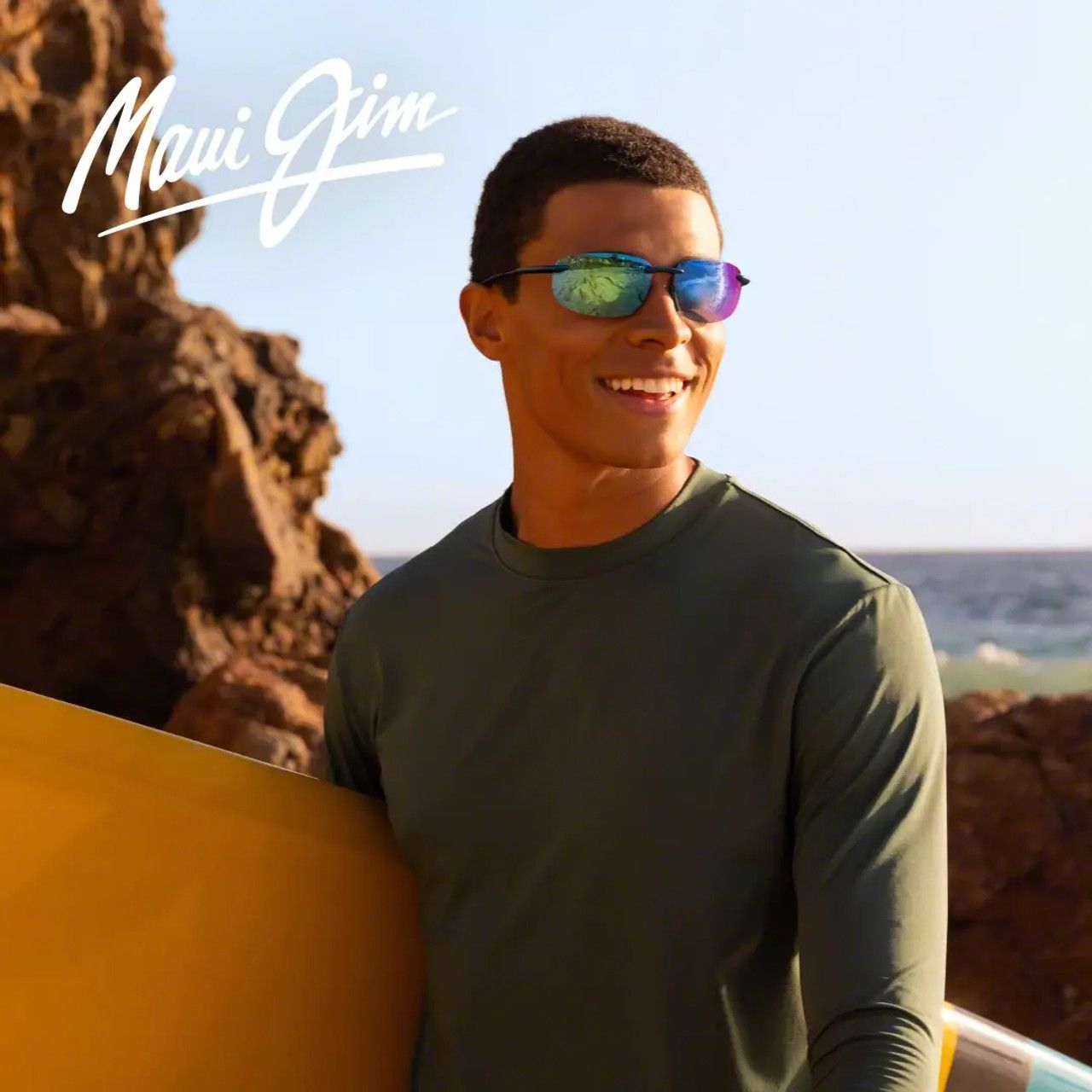Man wearing mirrored sunglasses holds a yellow surfboard on a sunny rocky beach, with “Maui Jim” logo