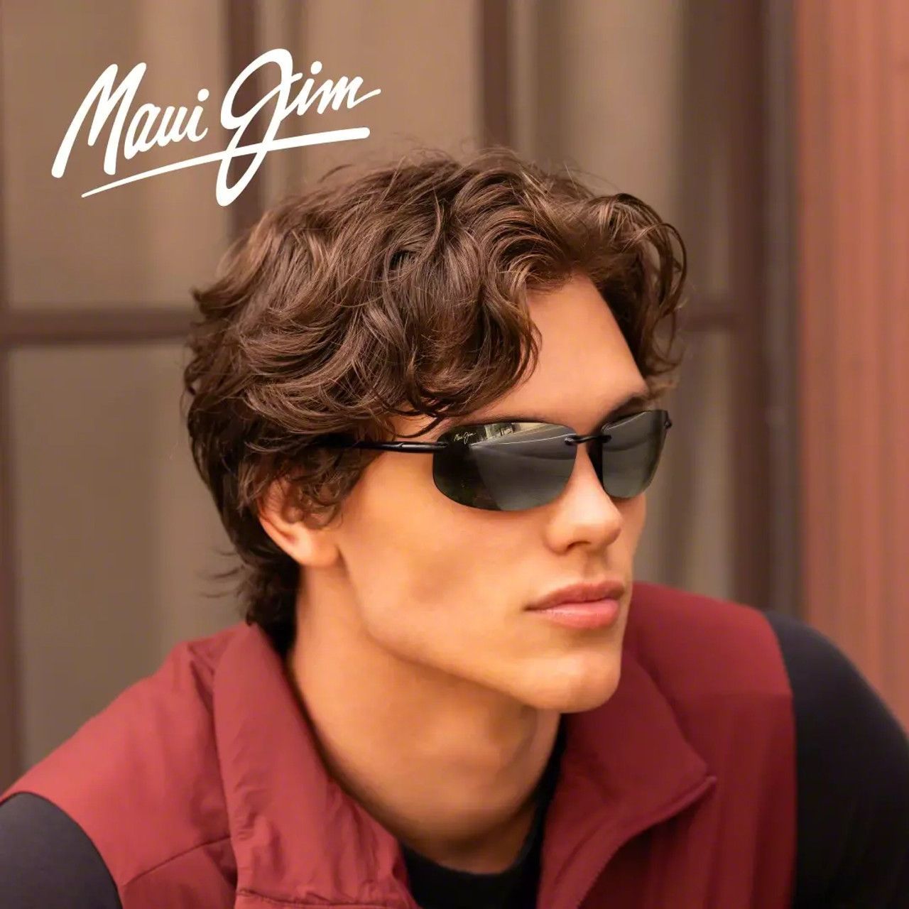 Man wearing sunglasses and a maroon jacket in a warm-toned portrait, with “Maui Jim” logo in the corner