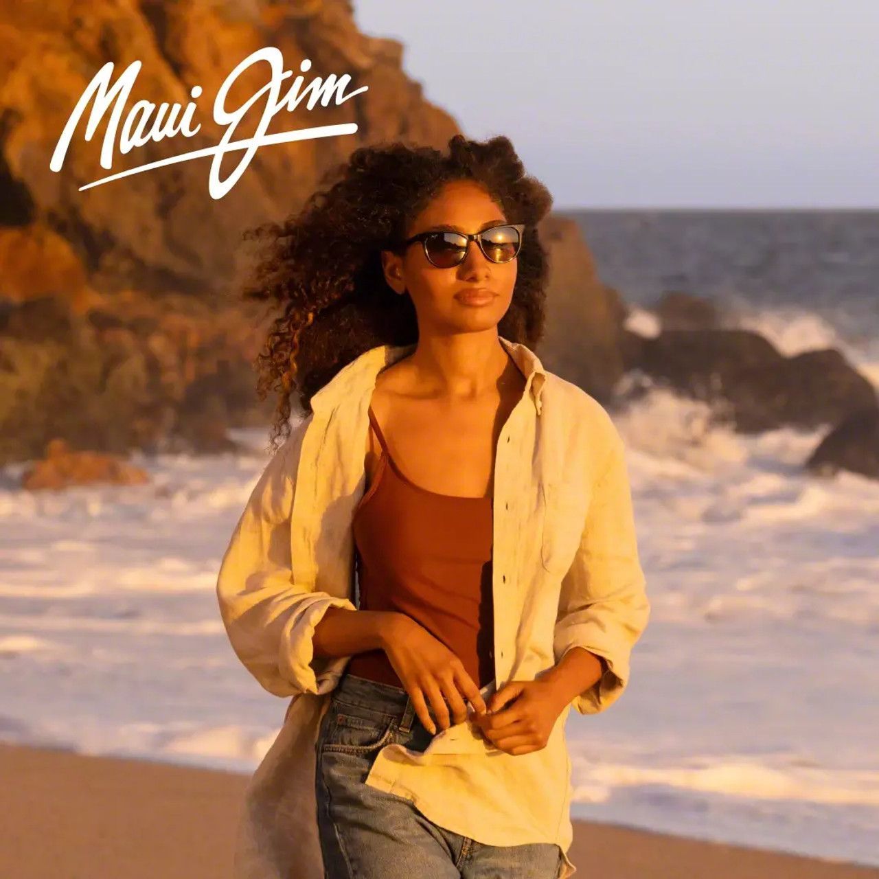 Woman in sunglasses and light shirt walking on a beach at sunset, with waves and Maui Jim logo.