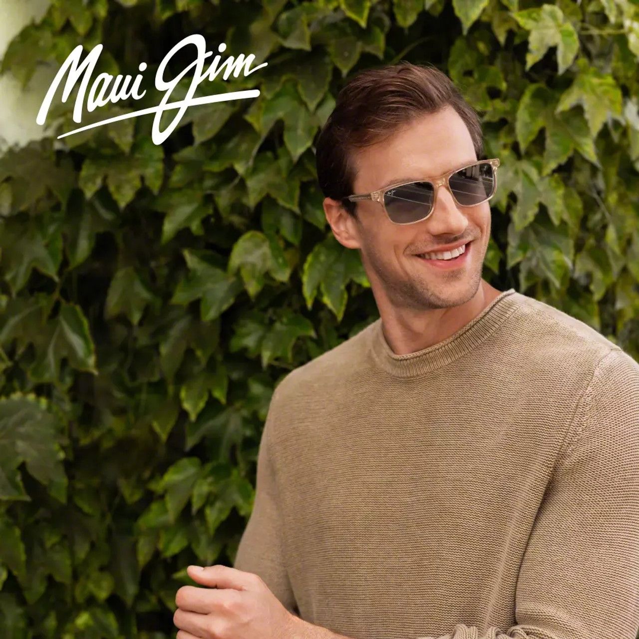 Smiling man in sunglasses and tan sweater standing before a leafy green wall