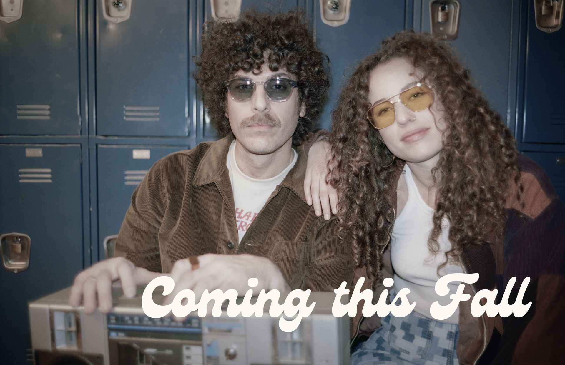 A man and a woman are sitting next to each other in front of lockers wearing moscot sunglasses.