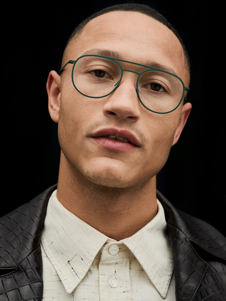 Portrait of a person wearing round glasses, a cream shirt, and a dark jacket against a black background