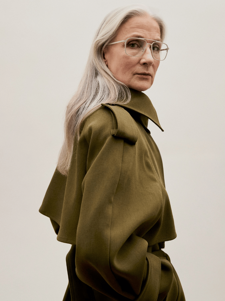 Portrait of a person with long silver hair and glasses wearing an olive coat against a plain background