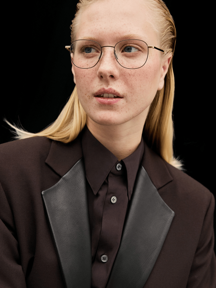 Blond person wearing round glasses and a dark suit against a black background.