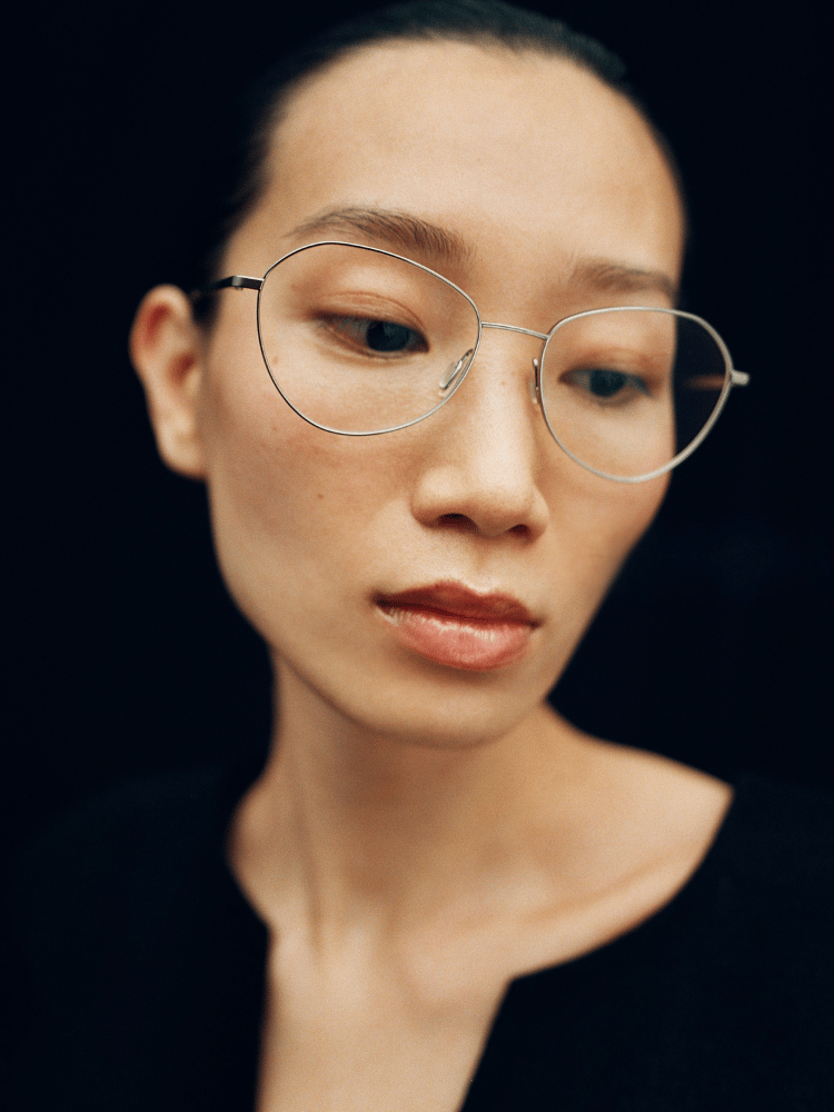 Portrait of a person wearing round glasses against a dark background, looking downward thoughtfully