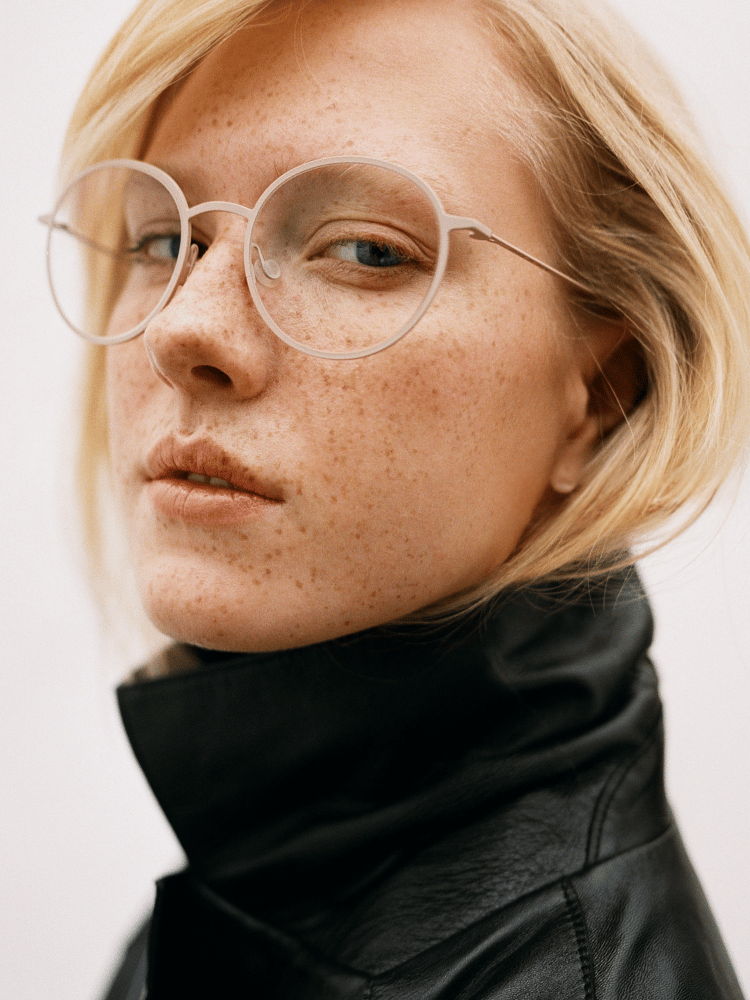 Portrait of a person with round glasses and freckles, wearing a black high-collar jacket