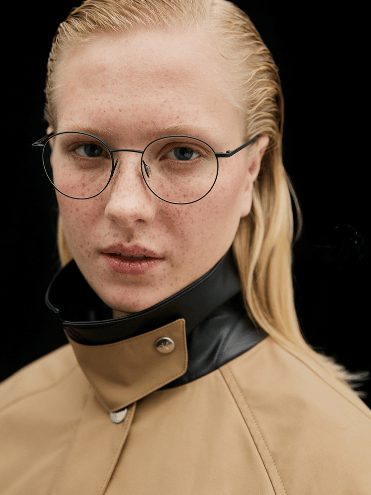 Portrait of a person in round glasses and a tan coat against a black background