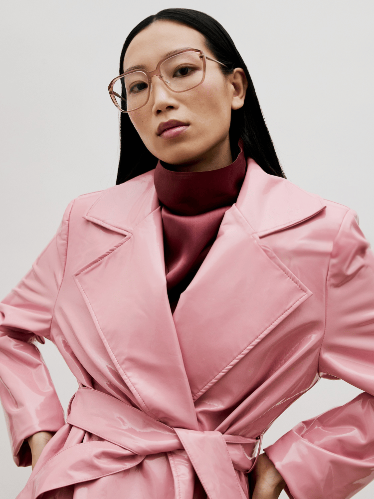 Person in a pink belted trench coat and glasses, posing against a plain light background.