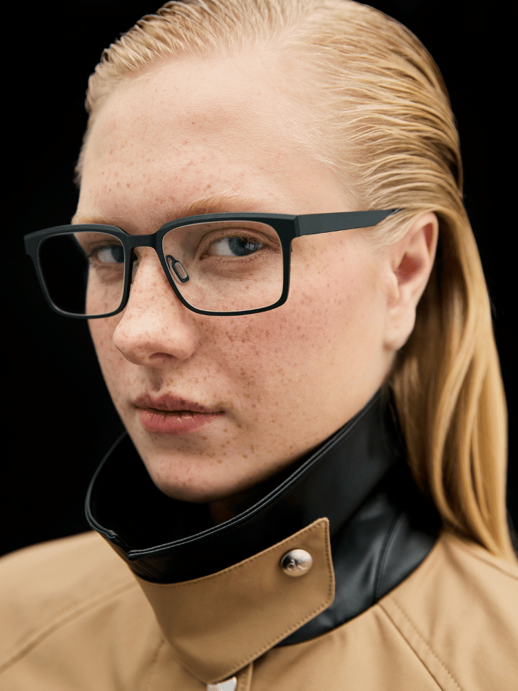 Person with slicked-back blond hair and black glasses wearing a tan-and-black high-collar jacket on a black background
