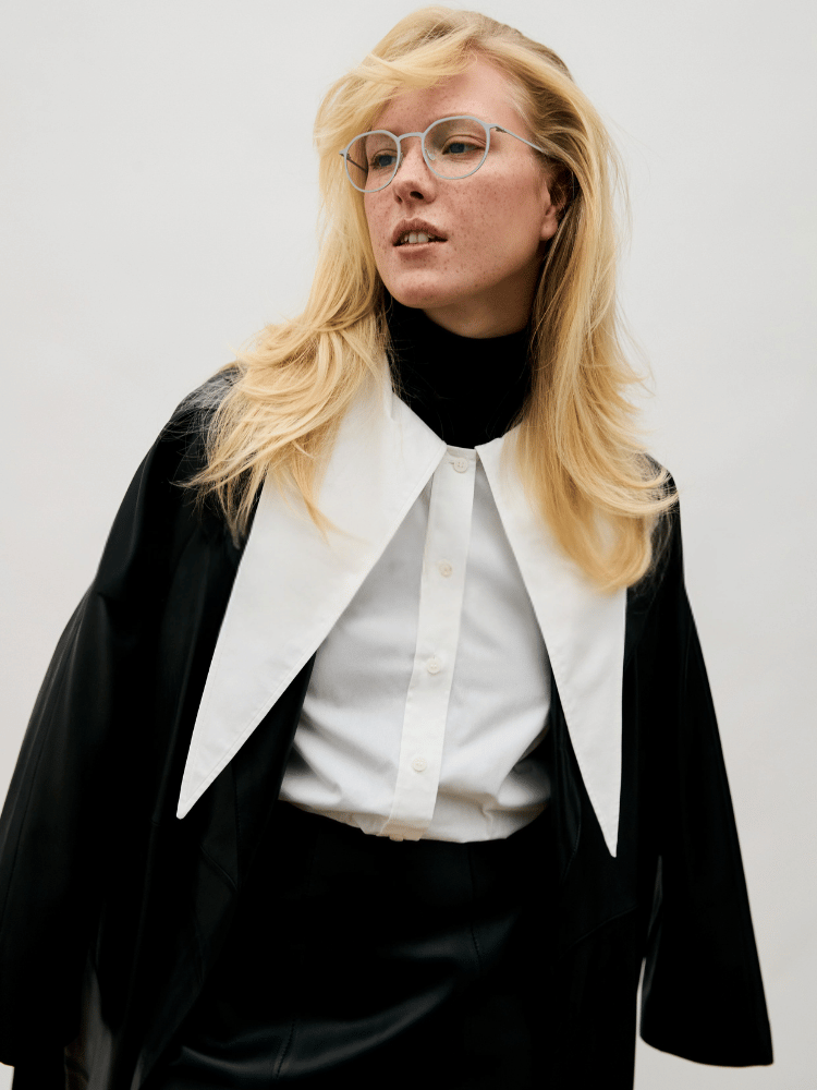 Blonde woman in glasses wearing a black cape and white shirt against a plain background