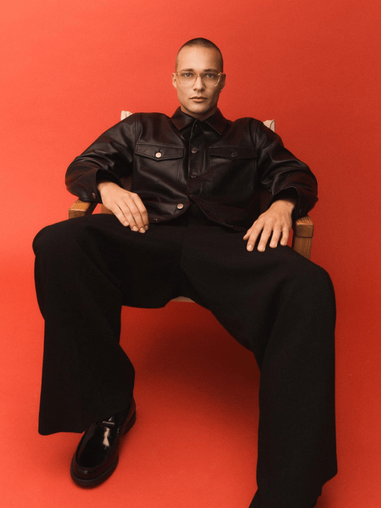A man is sitting in a chair wearing glasses, a black leather jacket and black pants.