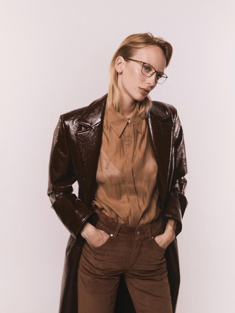 A woman wearing glasses and a brown coat is standing with her hands in her pockets.