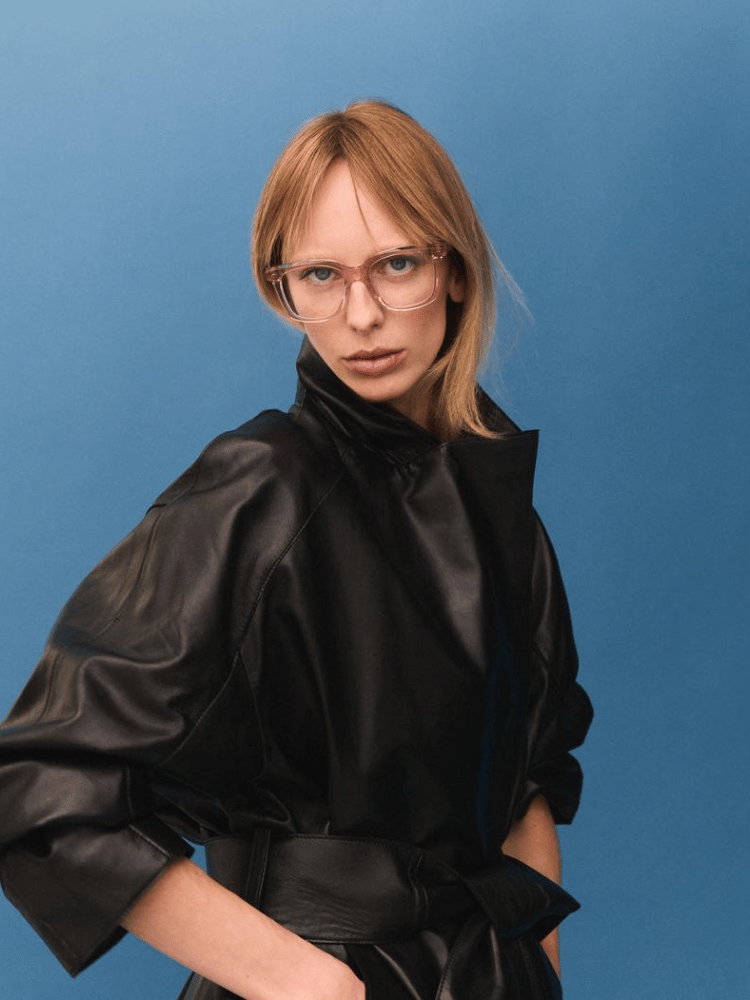 A woman wearing glasses and a black leather coat is standing in front of a blue background.