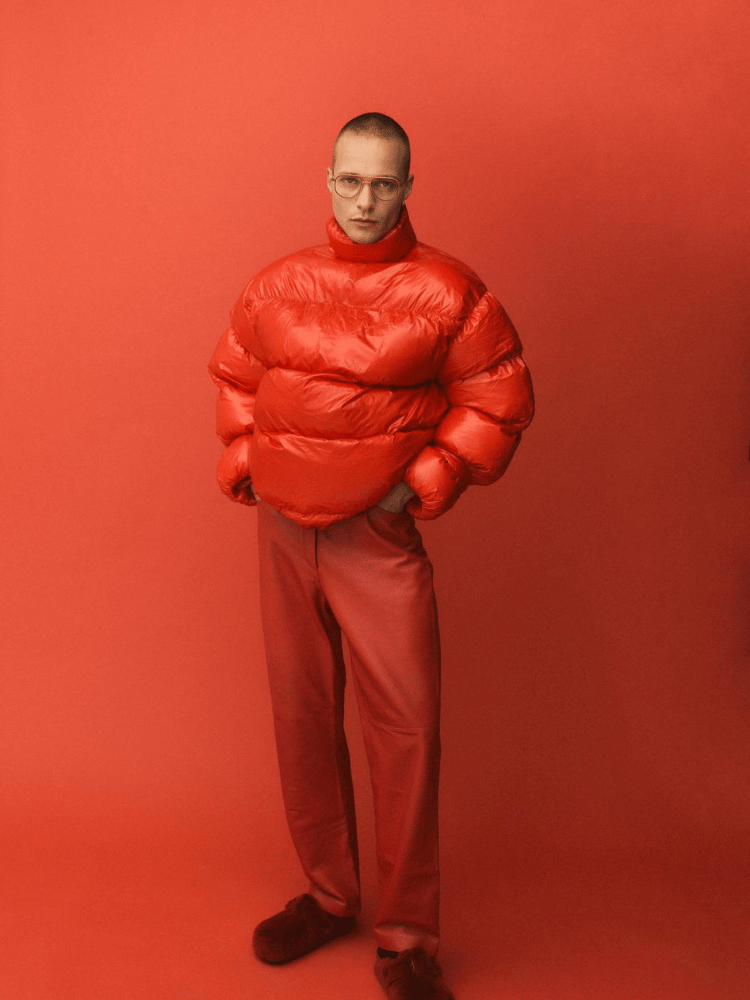 A man wearing glasses, a red jacket and red pants is standing in front of a red background.