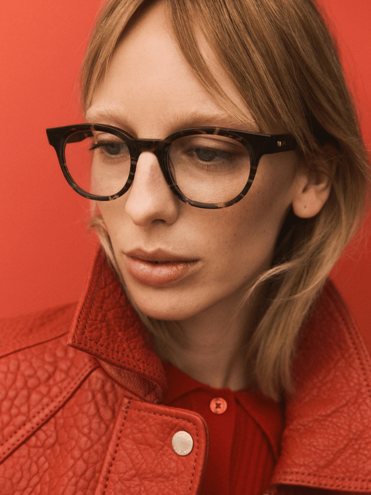 A woman wearing glasses and a red jacket is standing in front of a red wall.