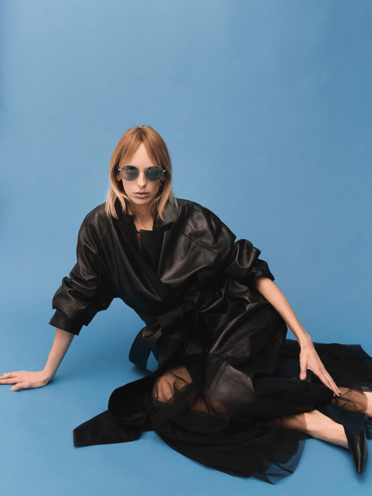 A woman in a black leather coat and sunglasses is sitting on the floor.