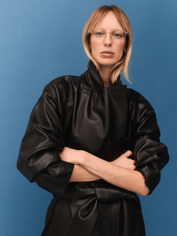 A woman wearing glasses and a black leather coat is standing with her arms crossed.