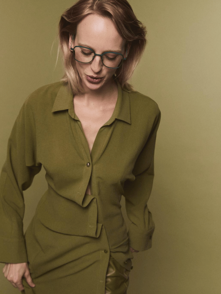 A woman wearing glasses and a green shirt is standing in front of a green background.
