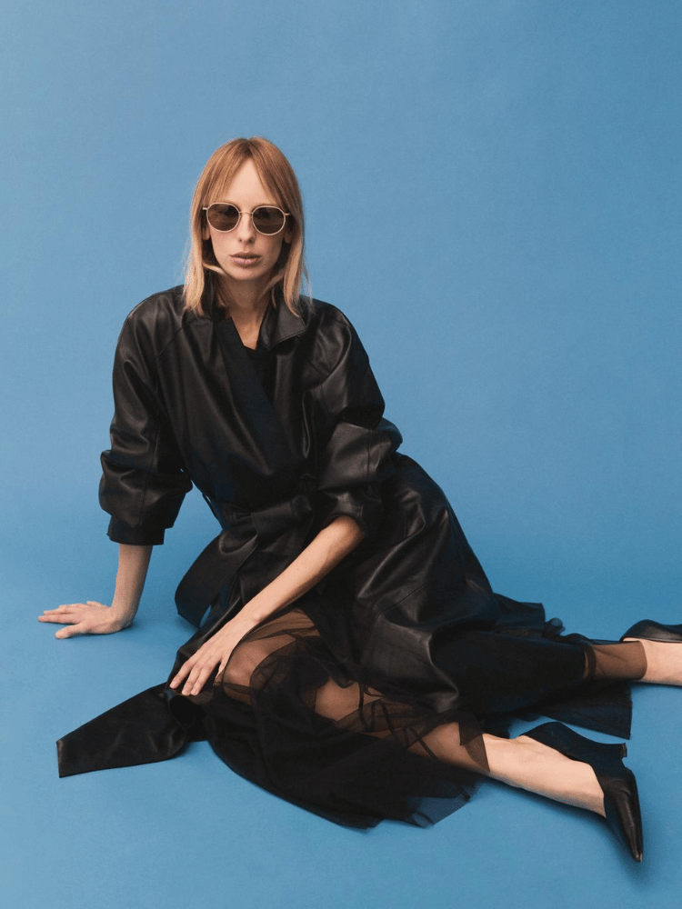 A woman wearing sunglasses, a black dress and leather jacket is sitting on the floor.