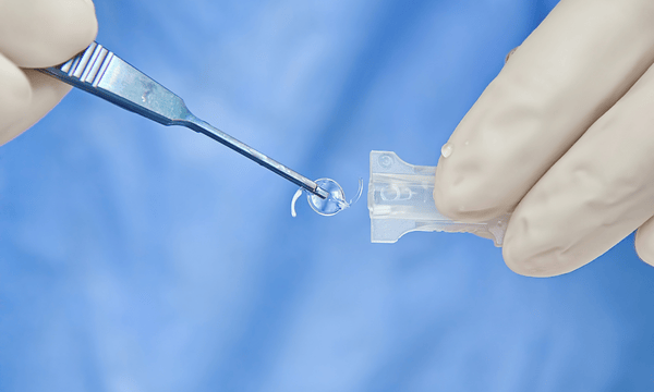 Surgical tools holding a clear, rolled-up lens next to a small plastic container, on a blue surgical drape.