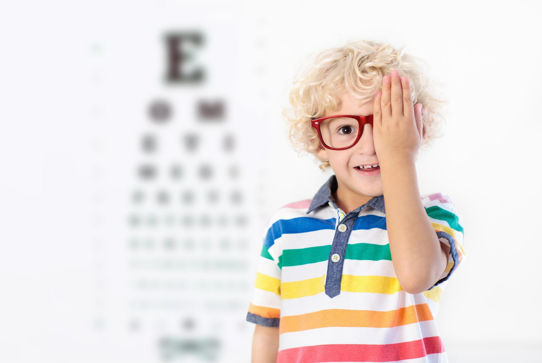 A young boy wearing glasses is covering his eye in front of an eye test chart.