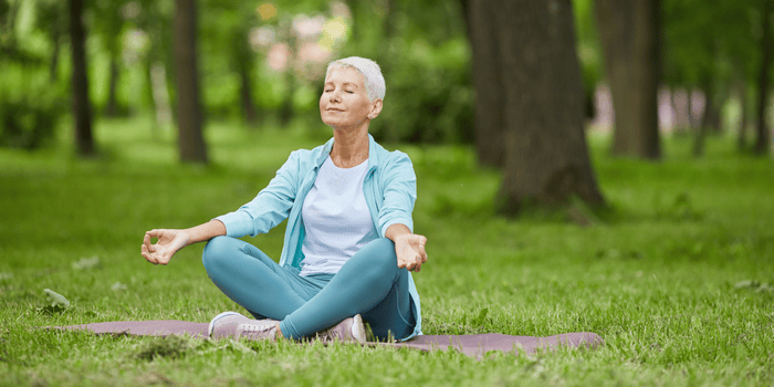 Woman in lotus position meditating in a park, eyes closed, surrounded by trees and grass.