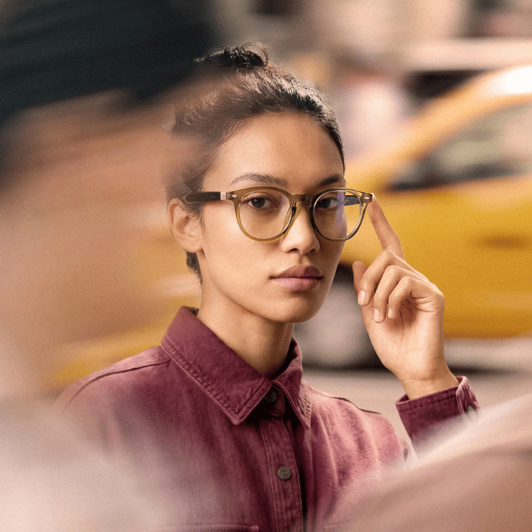 Woman in glasses and maroon shirt adjusting frames on a blurred city street
