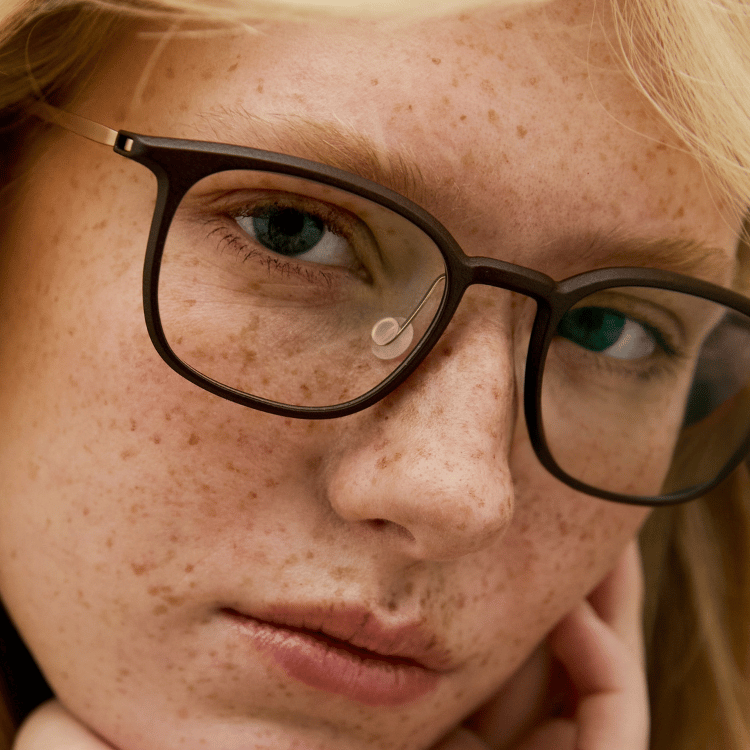 Close-up of a freckled person with blonde hair wearing dark glasses, looking calmly at the camera.