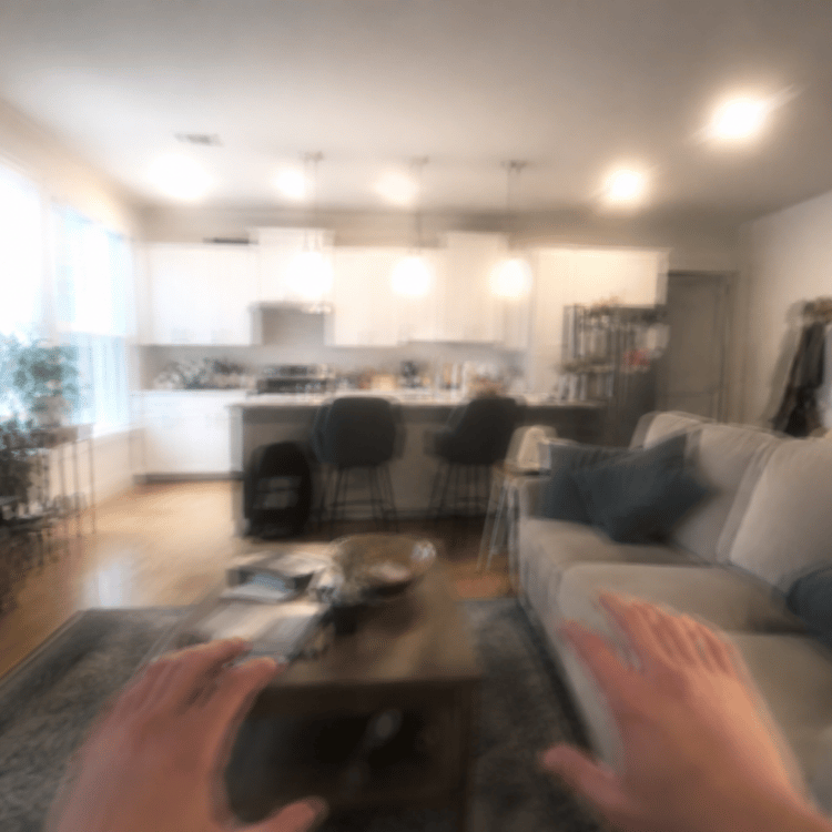 Blurry living room with kitchen, sofa, coffee table, and two hands visible in the foreground.