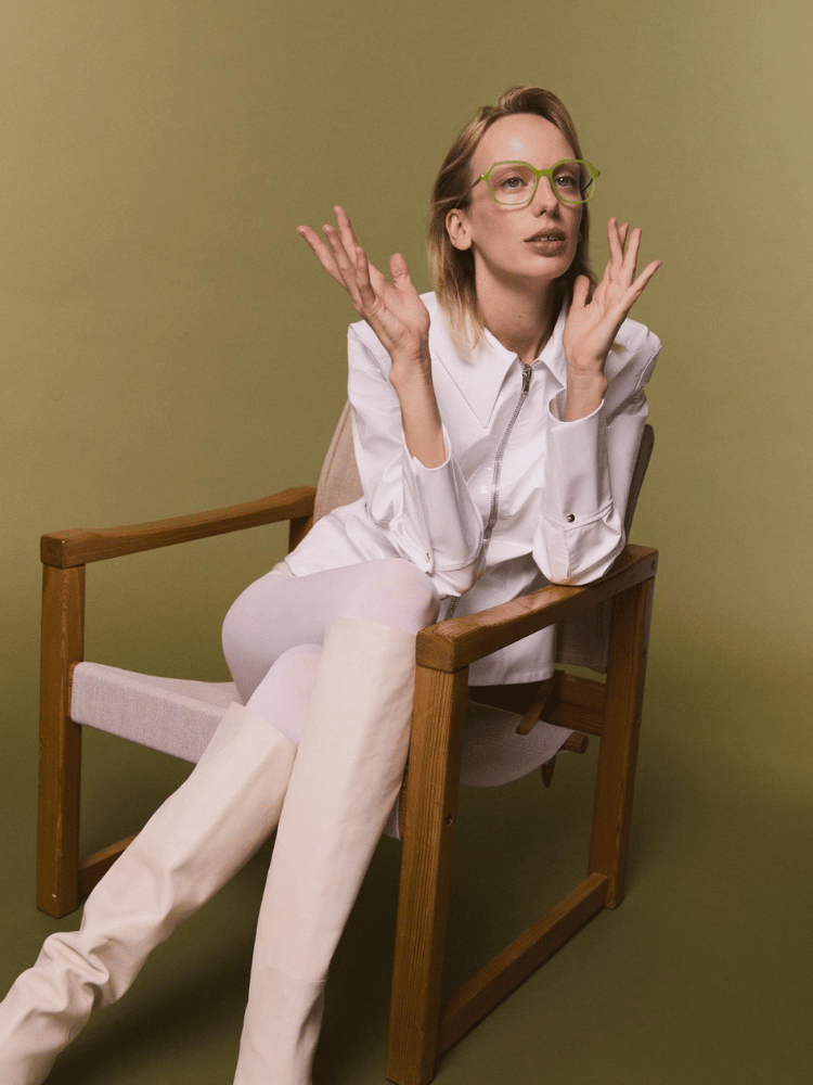 A woman wearing glasses , a white shirt and white boots is sitting in a chair.