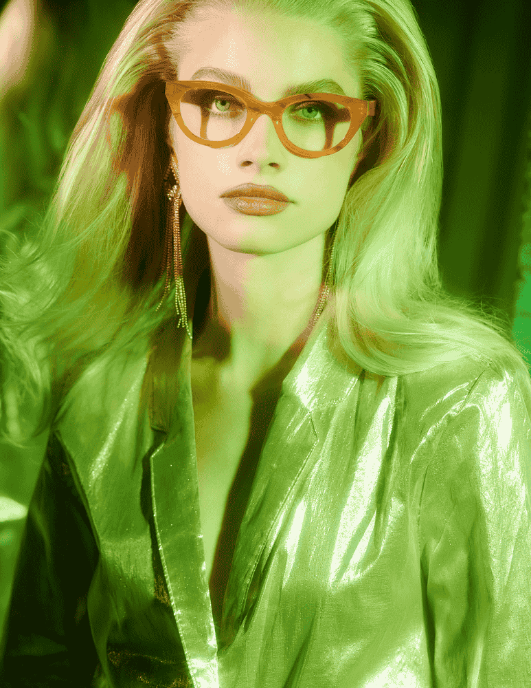 A woman wearing glasses and a green jacket is standing in front of a mirror.