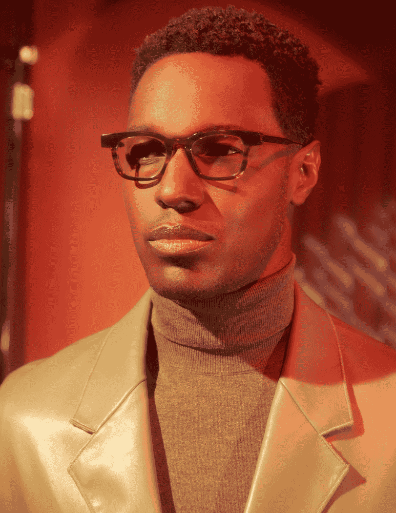 A man wearing glasses and a turtleneck is standing in front of a microphone.