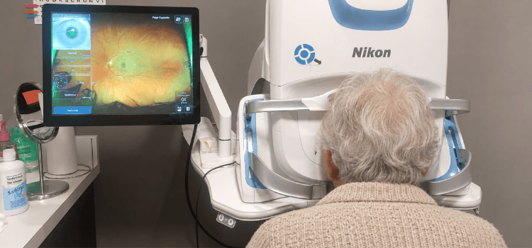Person seated at a Nikon eye scanner beside a monitor showing a colorful retinal image