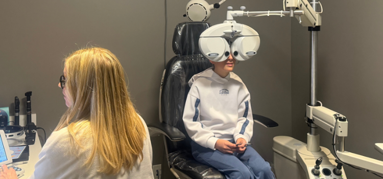 Person sitting for an eye exam in an optometry clinic, facing a phoropter with a doctor nearby.